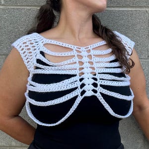 May include: A white crochet top featuring a rib cage design with openwork detailing. The garment is constructed with yarn in a skeletal pattern that covers the chest and shoulders, worn over a solid black base layer for high contrast.