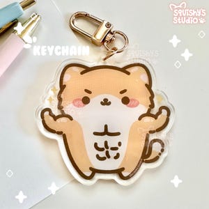 May include: A brown and white cartoon dog with a muscular body and a determined expression. The dog is wearing a gold keychain with a metal ring. The keychain is labeled "KEYCHAIN".