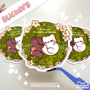 May include: A collection of stickers featuring a cute cartoon couple embracing. The stickers have a green background with white flowers and the text "I love you in every universe". The stickers are white and brown, with a heart-shaped design.
