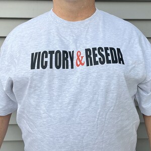 May include: A light gray heathered short-sleeve t-shirt featuring the text VICTORY & RESEDA printed across the chest. The word VICTORY and the word RESEDA are in black capital letters, while the ampersand symbol is in a bright orange color.