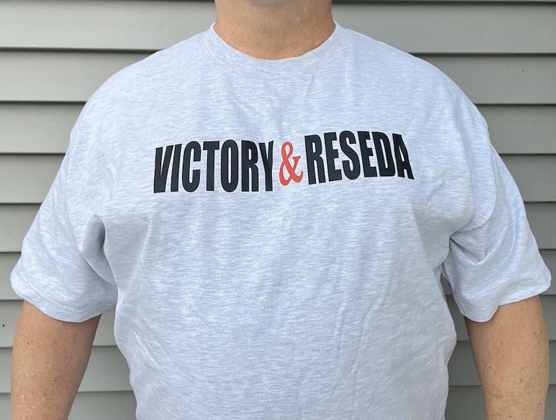 May include: A light gray heathered short-sleeve t-shirt featuring the text VICTORY & RESEDA printed across the chest. The word VICTORY and the word RESEDA are in black capital letters, while the ampersand symbol is in a bright orange color.