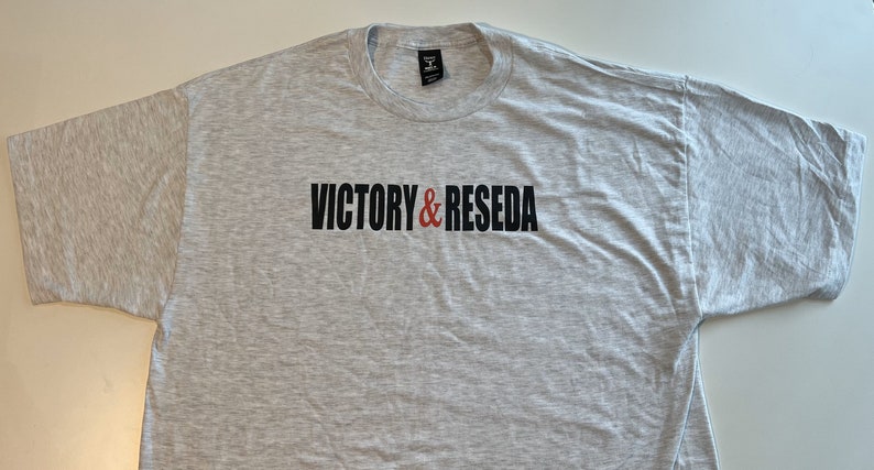 May include: A light gray heathered short-sleeve t-shirt featuring the text VICTORY & RESEDA printed in black and red on the chest. The garment has a crew neckline and a standard fit, with a black brand label visible inside the collar area.