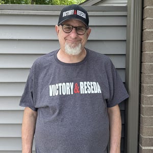 May include: A charcoal gray short-sleeved t-shirt featuring the white and red text VICTORY & RESEDA printed across the chest, paired with a matching black baseball cap that displays the same VICTORY & RESEDA text in white and red lettering.