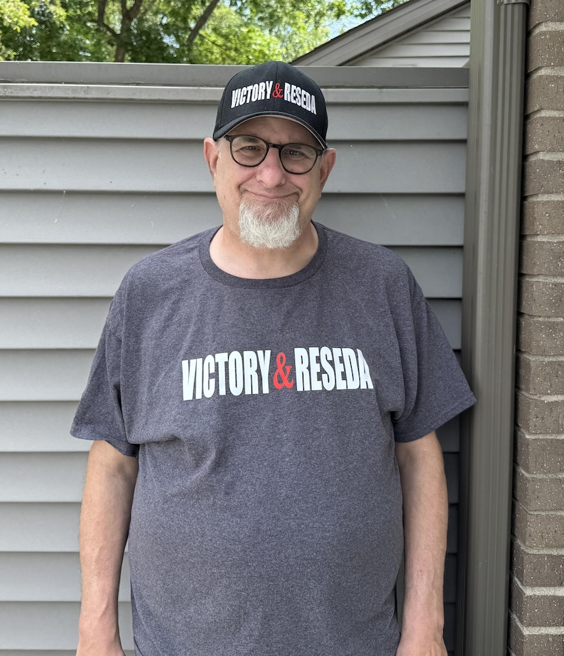 May include: A charcoal gray short-sleeved t-shirt featuring the white and red text VICTORY & RESEDA printed across the chest, paired with a matching black baseball cap that displays the same VICTORY & RESEDA text in white and red lettering.