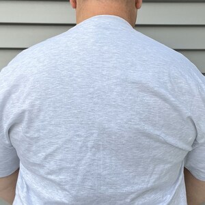 May include: A short-sleeved t-shirt in a light heather gray color. The garment features a crew neckline and a plain, solid-colored design with a textured, speckled appearance throughout the fabric, providing a simple and neutral look.