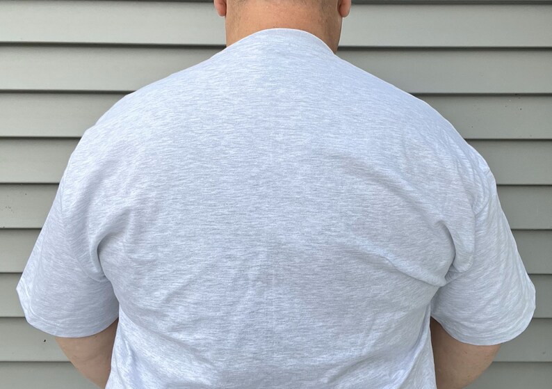 May include: A short-sleeved t-shirt in a light heather gray color. The garment features a crew neckline and a plain, solid-colored design with a textured, speckled appearance throughout the fabric, providing a simple and neutral look.