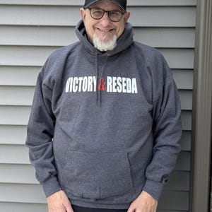 May include: A charcoal gray hooded sweatshirt featuring the white and red text VICTORY & RESEDA printed across the chest, paired with a matching black baseball cap that displays the same VICTORY & RESEDA text in white and red on the front.