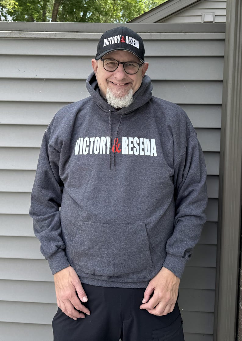 May include: A charcoal gray hooded sweatshirt featuring the white and red text VICTORY & RESEDA printed across the chest, paired with a matching black baseball cap that displays the same VICTORY & RESEDA text in white and red on the front.