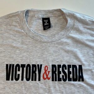 May include: A light gray heathered crew neck t-shirt featuring the text VICTORY & RESEDA printed across the chest. The word VICTORY and the word RESEDA are in black capital letters, while the ampersand symbol in the center is red.