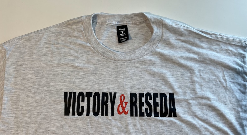 May include: A light gray heathered crew neck t-shirt featuring the text VICTORY & RESEDA printed across the chest. The word VICTORY and the word RESEDA are in black capital letters, while the ampersand symbol in the center is red.