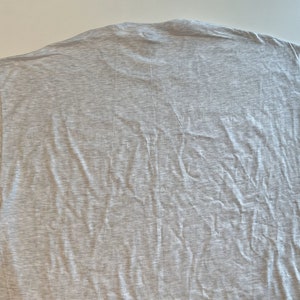 May include: A short-sleeved t-shirt in a light heather gray color. The garment features a crew neckline and a plain, unpatterned design. The fabric has a visible textured weave, and the shirt is laid out flat, showing the back side of the piece.