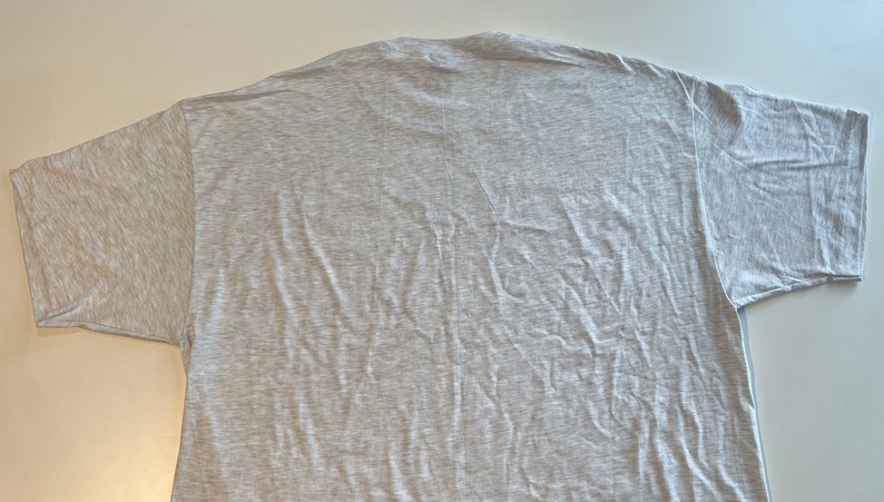 May include: A short-sleeved t-shirt in a light heather gray color. The garment features a crew neckline and a plain, unpatterned design. The fabric has a visible textured weave, and the shirt is laid out flat, showing the back side of the piece.