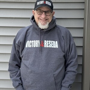 May include: A dark charcoal gray hooded sweatshirt featuring the white and red text VICTORY & RESEDA printed across the chest, paired with a matching black baseball cap that displays the same VICTORY & RESEDA text in white and red lettering.