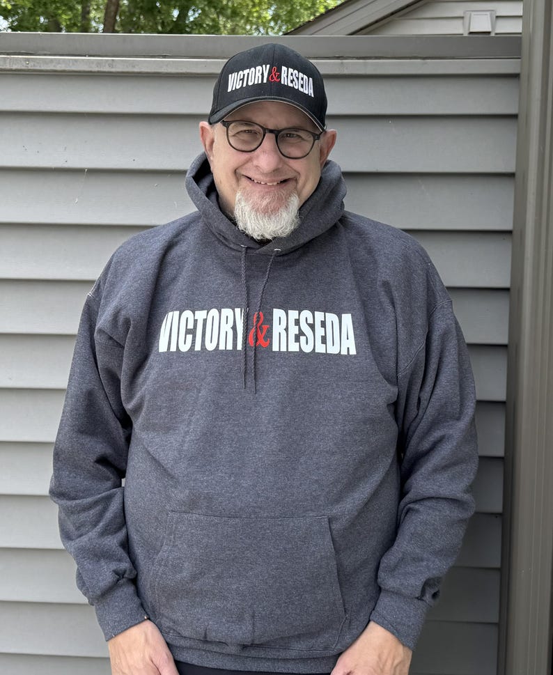 May include: A dark charcoal gray hooded sweatshirt featuring the white and red text VICTORY & RESEDA printed across the chest, paired with a matching black baseball cap that displays the same VICTORY & RESEDA text in white and red lettering.