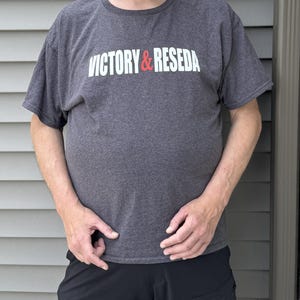May include: A charcoal grey short-sleeve t-shirt featuring the white and red text VICTORY & RESEDA across the chest, paired with a black baseball cap displaying the same text and black shorts with a side pocket and dark-rimmed eyeglasses.