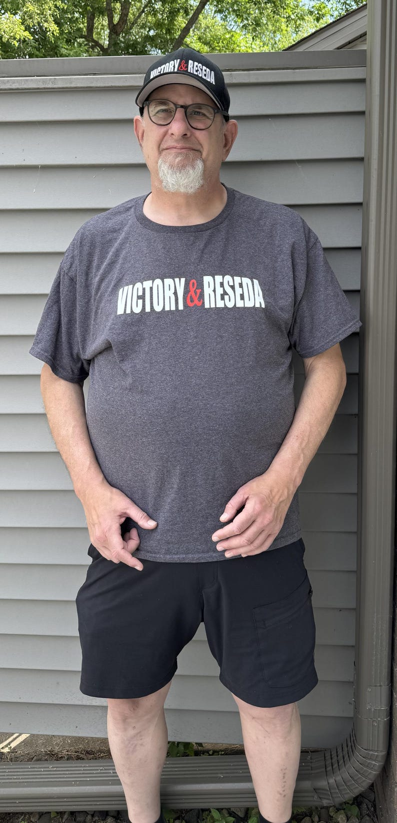 May include: A charcoal grey short-sleeve t-shirt featuring the white and red text VICTORY & RESEDA across the chest, paired with a black baseball cap displaying the same text and black shorts with a side pocket and dark-rimmed eyeglasses.