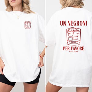 May include: White t-shirt with a red outline illustration of a cocktail glass on the front and back. The back of the shirt features the text "UN NEGRONI PER FAVORE" with "Florence, Italy 2019" below the text.