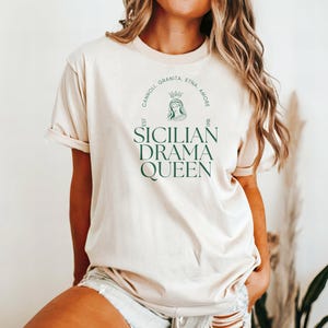 May include: A beige t-shirt with a green graphic design that says "Sicilian Drama Queen" with a circle around it that says "Cannoli, Granita, Etna, Amore" and "Est. 1861".