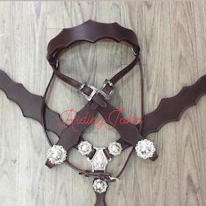 May include: A brown leather horse breast collar with decorative silver-tone conchos and buckles. The breast collar has a scalloped edge design and is laid out on a wooden surface.