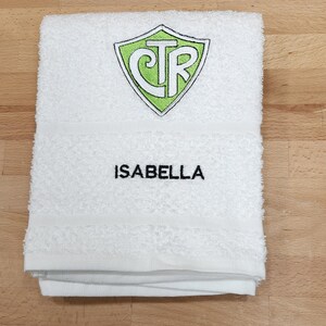 May include: White bath towel with the name "ISABELLA" embroidered in black. The towel features a green and white shield-shaped emblem with the letters "CTR". The towel is folded and placed on a wooden surface.
