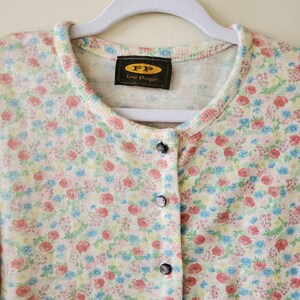 May include: A floral print cardigan with a round neckline and button-down front. The sweater features a pattern of small red, blue, and yellow flowers on a cream background. The label reads "FP Free People".