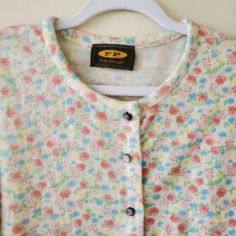 May include: A floral print cardigan with a round neckline and button-down front. The sweater features a pattern of small red, blue, and yellow flowers on a cream background. The label reads "FP Free People".