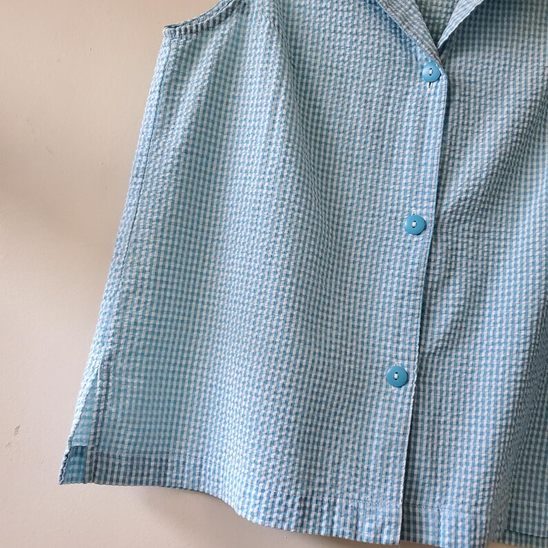 May include: A sleeveless, light blue and white seersucker shirt with a button-up front. The shirt has three light blue buttons and a classic collar. The fabric has a textured, checkered pattern.