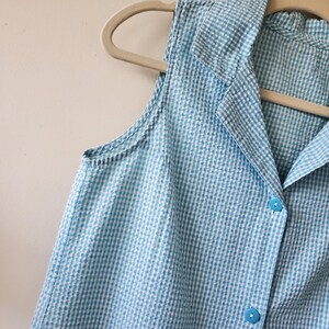 May include: A sleeveless, light blue and white gingham patterned top with a collared neckline and button-down front. The top is hanging on a beige hanger. The fabric has a textured appearance.