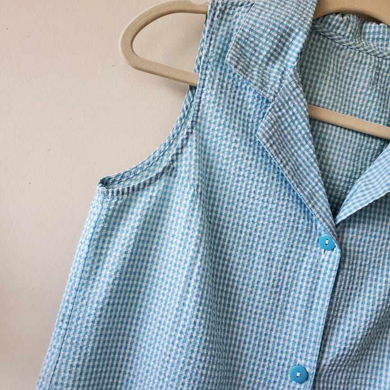 May include: A sleeveless, light blue and white gingham patterned top with a collared neckline and button-down front. The top is hanging on a beige hanger. The fabric has a textured appearance.