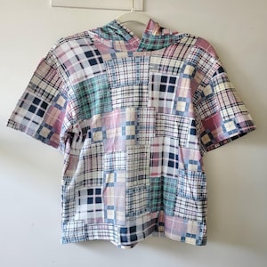 May include: A short-sleeved patchwork shirt with a hood. The shirt features a variety of plaid patterns in shades of blue, pink, green, and white. The shirt is hanging on a white hanger against a white wall.