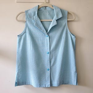 May include: A sleeveless, button-down shirt in a light blue and white checkered pattern. The shirt has a collared neckline and is hanging on a white hanger. The fabric has a textured appearance.