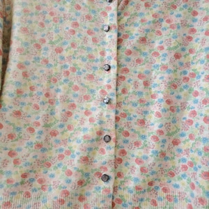 May include: A button-down cardigan sweater with a floral pattern in pink, blue, and yellow. The sweater has a ribbed hem and cuffs, and the buttons are a dark gray color. The background is a soft, off-white color.