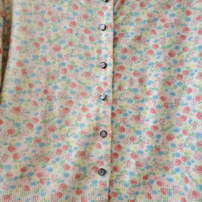 May include: A button-down cardigan sweater with a floral pattern in pink, blue, and yellow. The sweater has a ribbed hem and cuffs, and the buttons are a dark gray color. The background is a soft, off-white color.