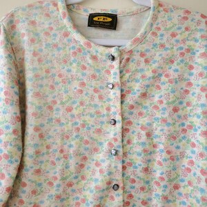 May include: A cream-colored cardigan sweater with a floral pattern in pink, blue, and yellow. The sweater has a round neckline, long sleeves, and a button-down front. The label reads "FP Free People".