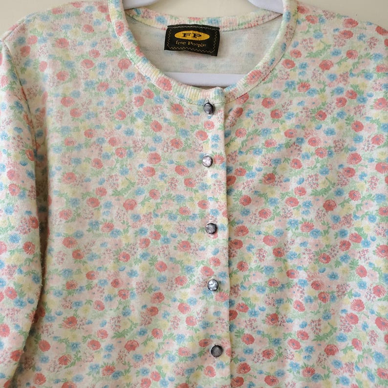 May include: A cream-colored cardigan sweater with a floral pattern in pink, blue, and yellow. The sweater has a round neckline, long sleeves, and a button-down front. The label reads "FP Free People".