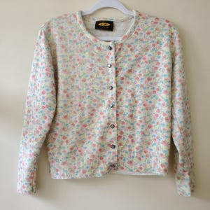May include: A floral print cardigan in pastel colors of pink, blue, and yellow. The button-down sweater has a round neckline and long sleeves. The garment is hanging on a white hanger. The label reads "FP Free People".