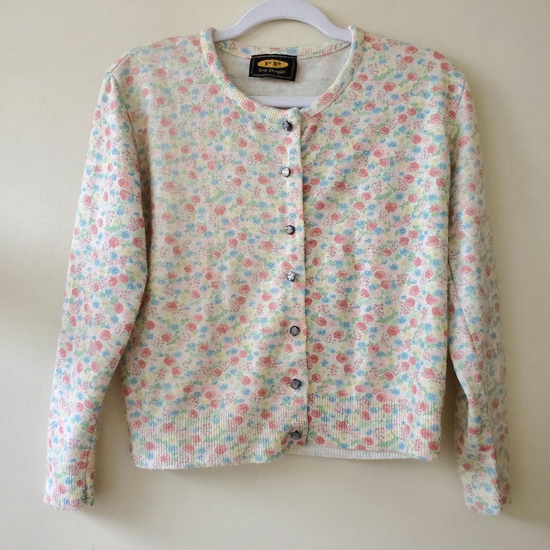May include: A floral print cardigan in pastel colors of pink, blue, and yellow. The button-down sweater has a round neckline and long sleeves. The garment is hanging on a white hanger. The label reads "FP Free People".