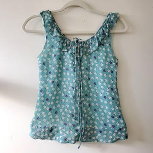 May include: A turquoise sleeveless top with a white and purple polka dot pattern. It has a ruffled neckline and a drawstring. The top is hanging on a white hanger.
