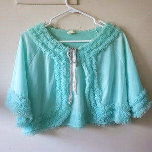 Könnte beinhalten: Ein vintage-farbenes, aquablaues Bolero-Jäckchen mit Rüschenbesatz an Kragen und Ärmeln. Die Jacke hat einen Verschluss mit silbernem Satinband. Das Kleidungsstück hängt an einem weißen Kleiderbügel.
