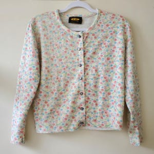 May include: A light-colored cardigan sweater with a floral pattern in pastel colors. The sweater has a round neckline, long sleeves, and a button-down front. The floral pattern includes pink, blue, and yellow flowers.