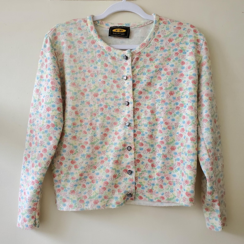 May include: A light-colored cardigan sweater with a floral pattern in pastel colors. The sweater has a round neckline, long sleeves, and a button-down front. The floral pattern includes pink, blue, and yellow flowers.