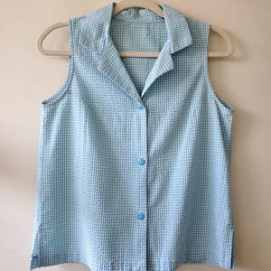 May include: A sleeveless, button-down shirt in a light blue and white gingham pattern. The shirt has a collared neckline and is hanging on a beige hanger. The fabric has a textured appearance.