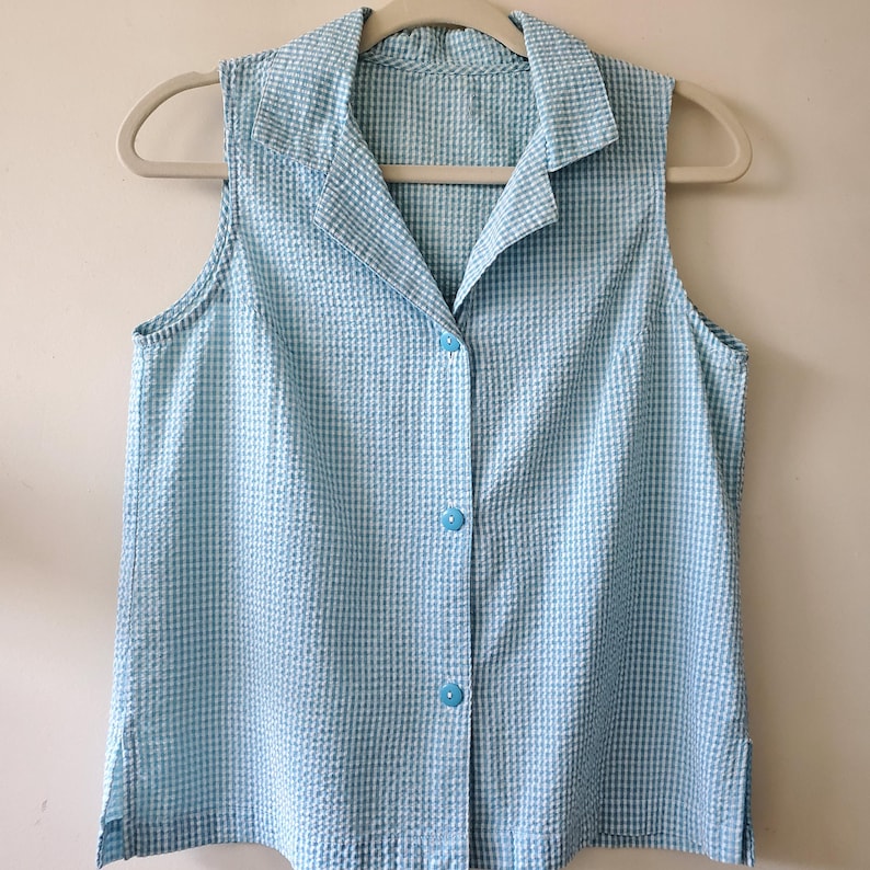 May include: A sleeveless, button-down shirt in a light blue and white gingham pattern. The shirt has a collared neckline and is hanging on a beige hanger. The fabric has a textured appearance.