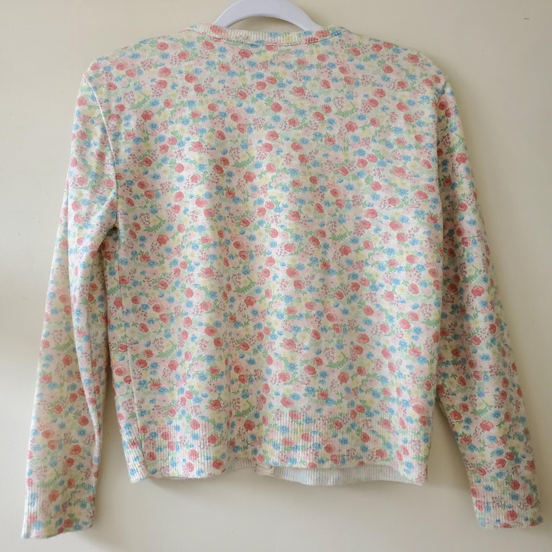 May include: A cream-colored cardigan sweater with a delicate floral pattern in shades of pink, blue, and yellow. The long-sleeved sweater is hanging on a white hanger, showcasing its soft, knit texture and classic design.