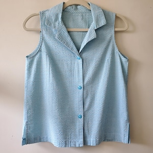 May include: A sleeveless, button-down shirt in a light blue and white checkered pattern. The shirt has a collared neckline and is hanging on a beige hanger. The buttons are a matching light blue color.