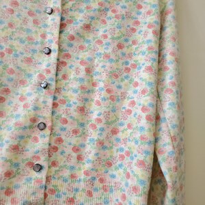 May include: A light-colored cardigan sweater with a floral pattern in pink, blue, and yellow. The sweater has a button-up front with silver buttons. The floral design is spread across the entire sweater.