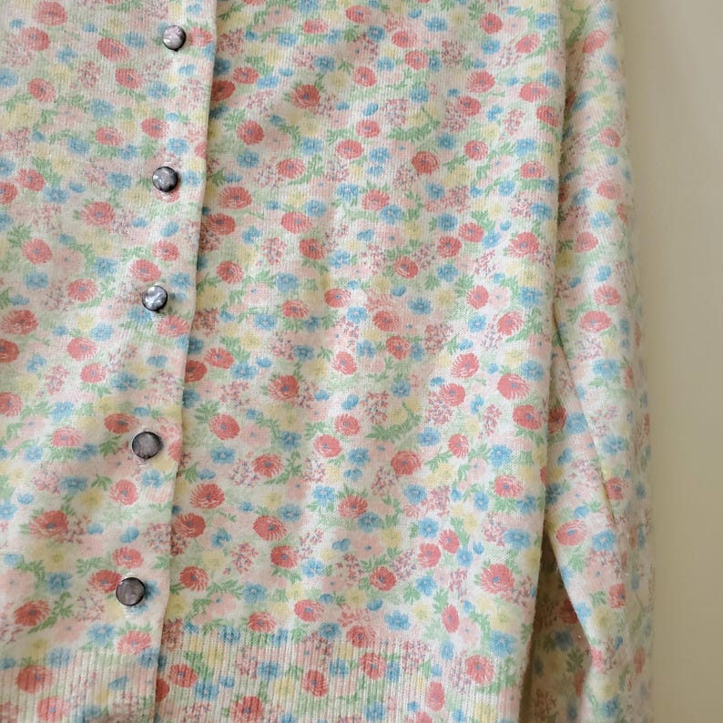 May include: A light-colored cardigan sweater with a floral pattern in pink, blue, and yellow. The sweater has a button-up front with silver buttons. The floral design is spread across the entire sweater.