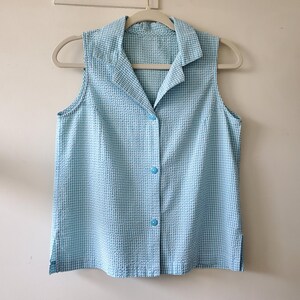 May include: A sleeveless, light blue and white checkered button-down shirt with a collar. The shirt is hanging on a gray hanger against a white wall. The shirt has three blue buttons down the front.