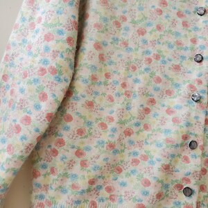 May include: A cream-colored cardigan sweater with a floral pattern in shades of pink, blue, and yellow. The sweater has a button-up front with four round, iridescent buttons. The sleeves are long, and the fabric appears soft and textured.
