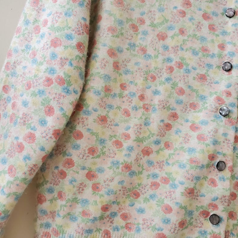 May include: A cream-colored cardigan sweater with a floral pattern in shades of pink, blue, and yellow. The sweater has a button-up front with four round, iridescent buttons. The sleeves are long, and the fabric appears soft and textured.